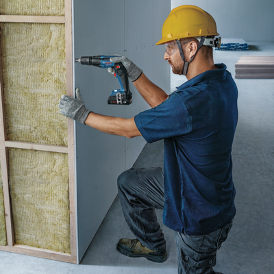 Trabalhador com capacete a usar furadeira elétrica para fixar placa de gesso em estrutura de madeira com isolamento.