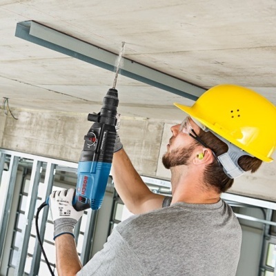 Homem com capacete amarelo furando teto de cimento com furadeira Bosch azul e preta