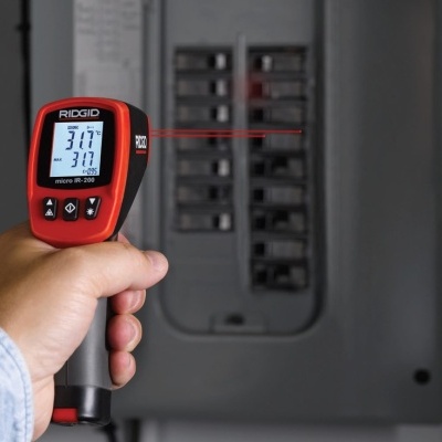 Termómetro infravermelho RIDGID vermelho e preto a medir temperatura num quadro elétrico cinza