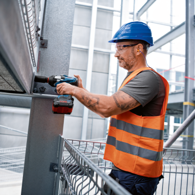 Homem com capacete azul e colete refletor a usar furadeira num ambiente industrial