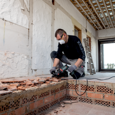 Homem a usar máscara e luvas a demolir tijolos vermelhos com martelo elétrico Bosch numa construção interior