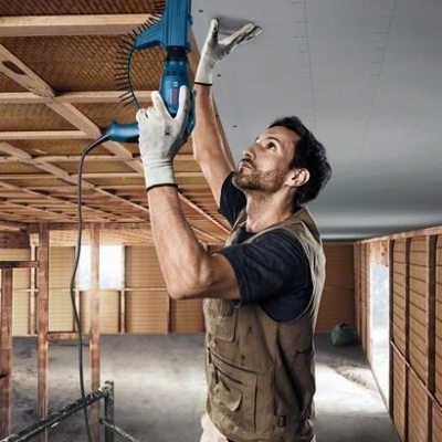 Homem a usar ferramenta elétrica azul para fixar material no teto de construção interior em madeira.