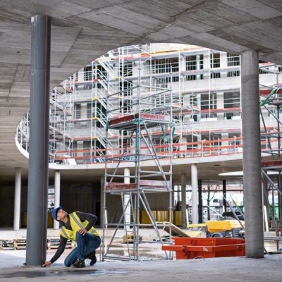 Área de construção com pilares de betão, andaimes e trabalhador em colete e capacete.