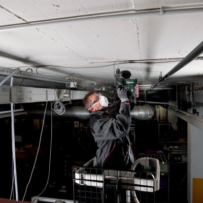 Homem a usar furadeira elétrica no teto de um espaço industrial.