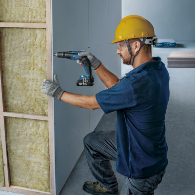 Homem com capacete amarelo a usar furadeira para fixar painel de gesso em obra