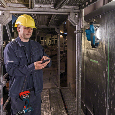 Homem com capacete amarelo e roupa de trabalho azul utilizando smartphone perto de andaime metálico e luz de trabalho azul fixa na parede