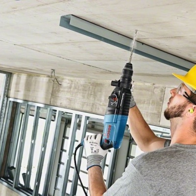 Homem com capacete amarelo a usar furadeira no teto de concreto numa construção