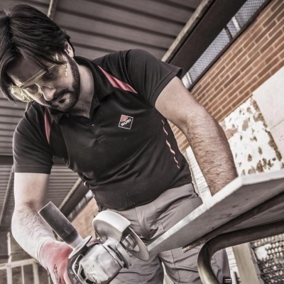 Homem a usar esmeril angular numa peça de metal, vestido com camisa preta e óculos protectores, em zona exterior coberta