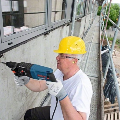 Homem a usar capacete amarelo e furadeira Bosch em obra na parede de edifício