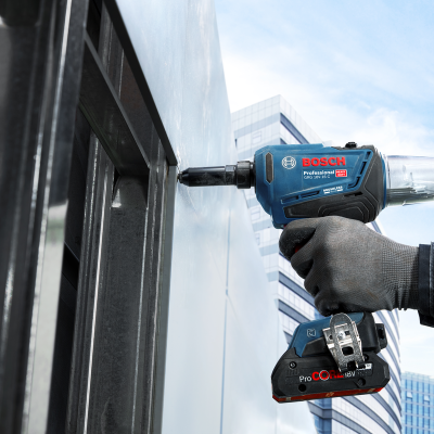 Parafusadora Bosch azul escura com mão enluvada a fixar peça metálica cinzenta ao vivo e fundo de edifícios