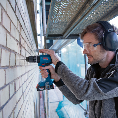 Homem a usar furadeira Bosch numa parede de tijolos clara com proteção auricular e óculos