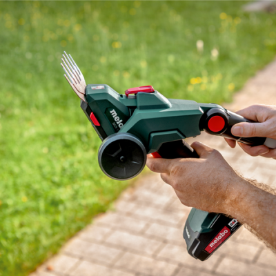 Aparelho de jardinagem Metabo verde com lâminas metálicas e rodas pretas em mãos sobre relva e pavimento