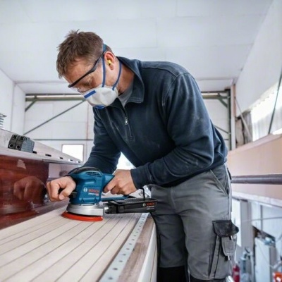 Homem a lixar madeira com lixadeira elétrica Bosch azul, com equipamento de proteção