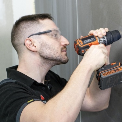 Homem a usar furadeira elétrica preto e laranja numa parede cinzenta.