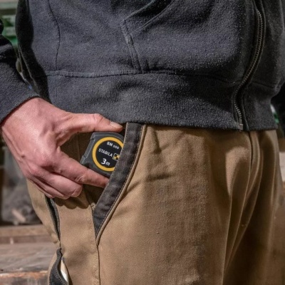 Pessoa a guardar fita métrica preta e amarela no bolso da calça bege numa oficina