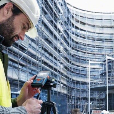 Homem com capacete e colete de segurança usando aparelho eletrônico junto de construção