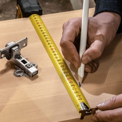 Mãos a medir e marcar madeira clara com fita métrica amarela, lápis branco e dobradiça metálica ao lado