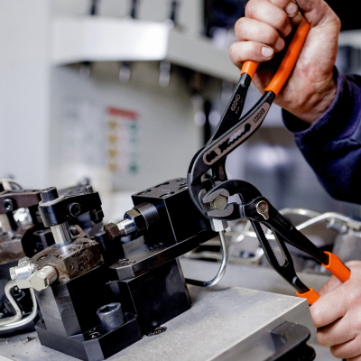 Alicates preto e laranja usados para segurar peças metálicas numa máquina industrial