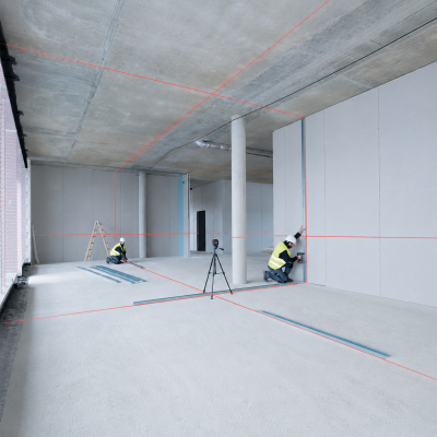 Espaço interno em construção com paredes cinza claras, colunas brancas, dois operários e linhas laser vermelhas