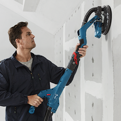Homem a lixar uma parede branca interior com lixadora eléctrica azul e preta