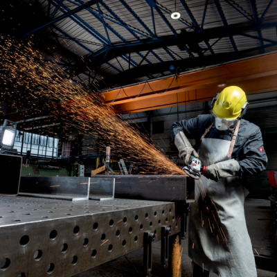 Trabalhador com capacete amarelo e avental cinza a usar rebarbadora em mesa metálica em oficina industrial