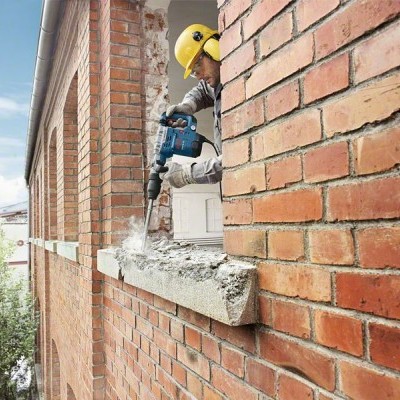 trabalhador com capacete amarelo a perfurar parapeito de cimento numa parede de tijolos vermelhos