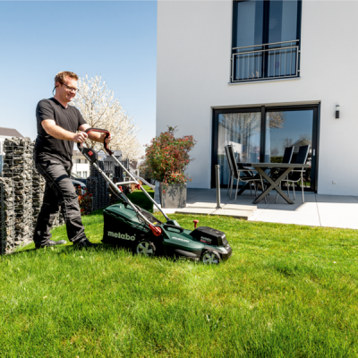 Cortador de relva Metabo verde escuro a ser usado num jardim em frente a casa moderna branca