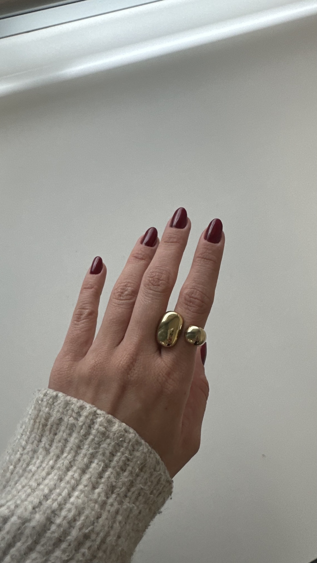 Carlota Ring Mão com anel dourado e unhas vinho sobre fundo branco.