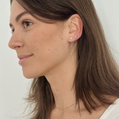 Mulher com três brincos pequenos dourados na orelha esquerda e colar fino dourado.