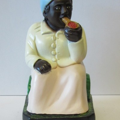 Figura decorativa de mulher sentada com casaco bege e lenço azul fumando cachimbo