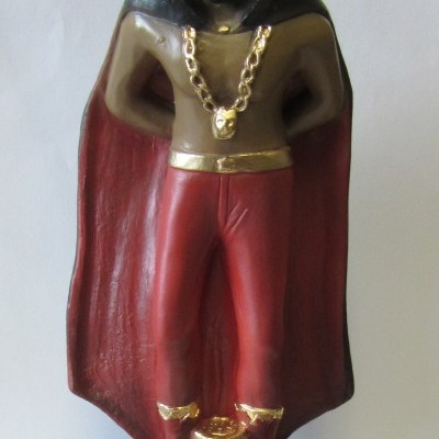 Escultura de homem vestido com chapéu, capa, calças vermelhas e acessórios dourados numa base preta