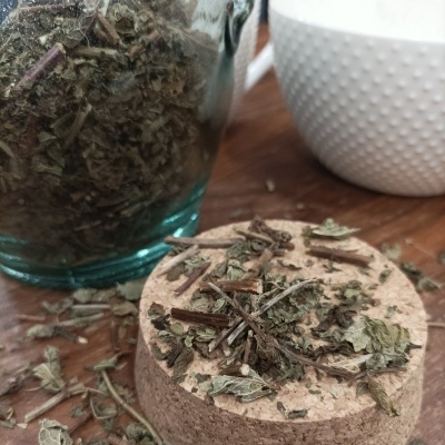 Frasco de vidro com ervas secas, tampo de cortiça e copo branco texturizado numa mesa de madeira.