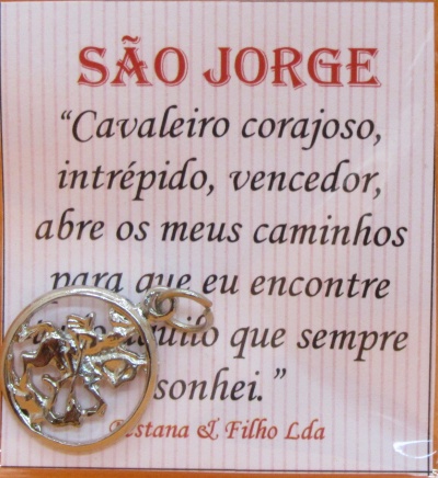 Pendente prateado de São Jorge com cartão de fundo rosa claro e texto em português.