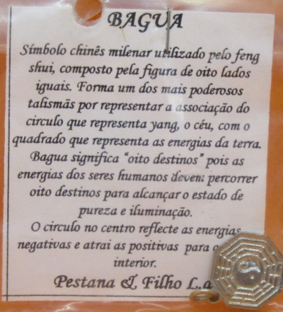Etiqueta com texto sobre o símbolo chinês Bagua e uma figura metálica do símbolo em baixo à direita.