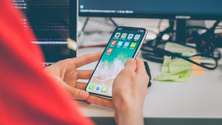 Pessoa a segurar telemóvel com ecrã ligado, mesa branca, papéis coloridos e cabos pretos ao fundo