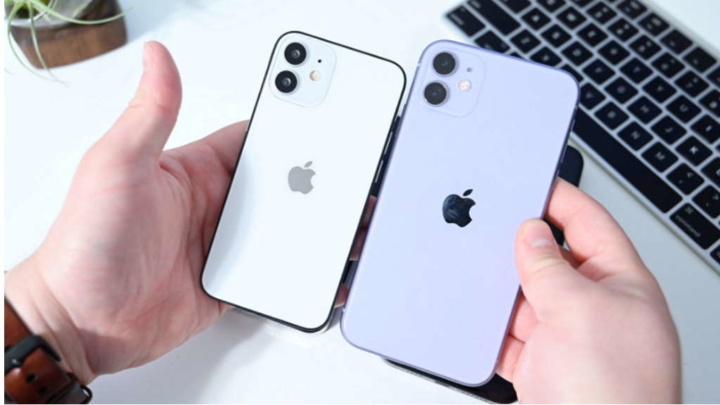 Duas mãos seguram dois iPhones, um branco e um lilás, com teclado ao fundo