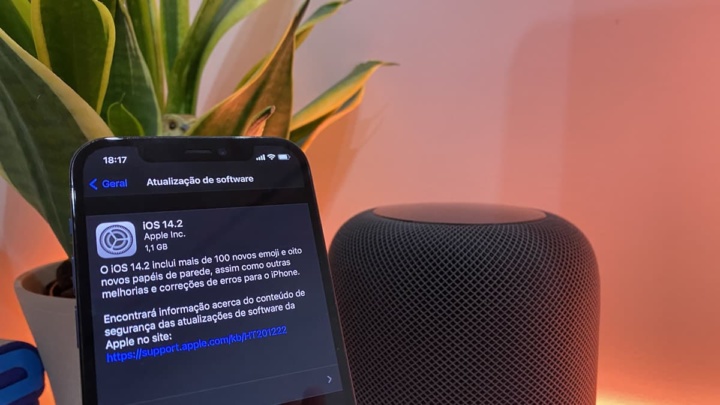 Smartphone preto com atualização iOS 14.2 ao lado de coluna de som cinza escura com plantas ao fundo