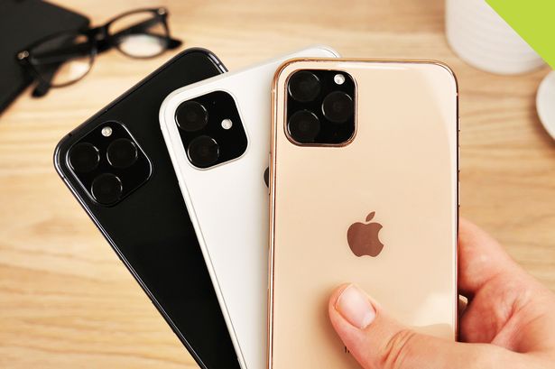 Três smartphones estilo iPhone em preto, branco e dourado sobre mesa de madeira