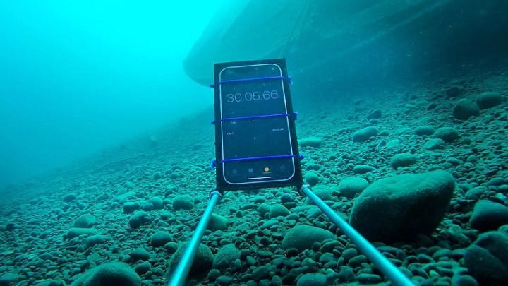Smartphone com cronómetro submerso sobre pedras com barco ao fundo.