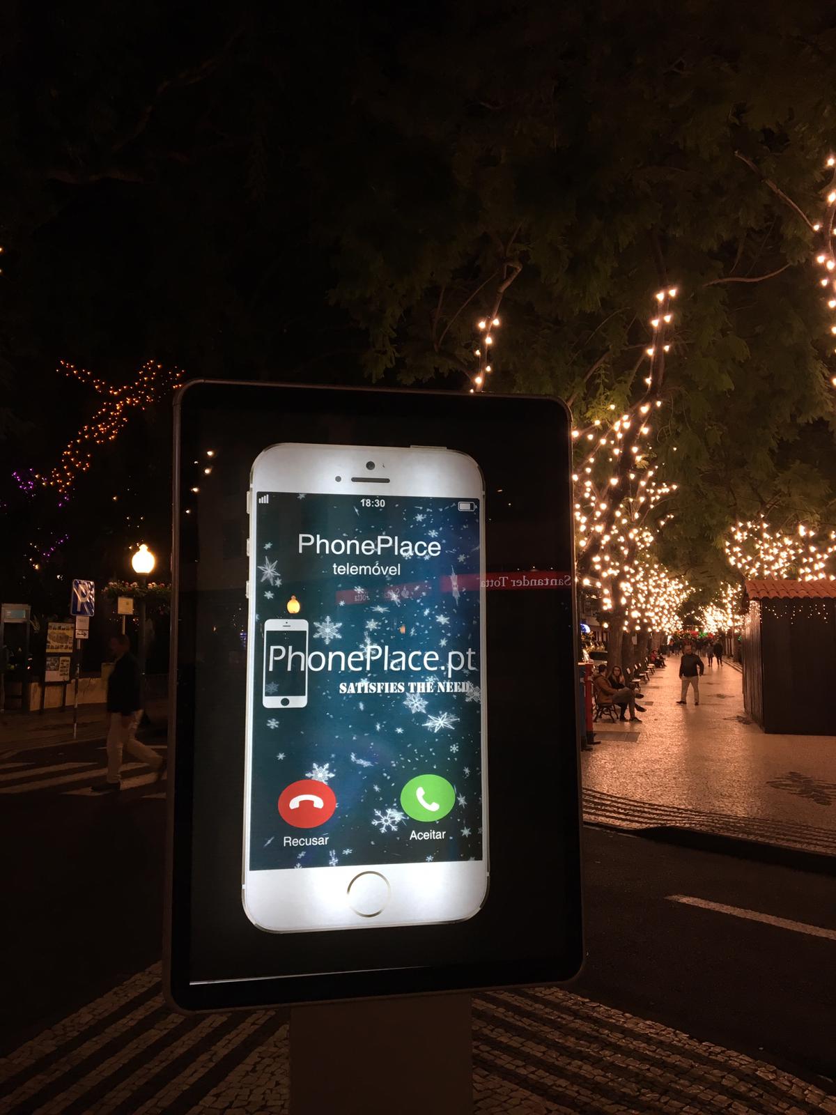 Receba o seu código de desconto de 10€ para as Prendas de Natal! Painel digital com anúncio de smartphone PhonePlace iluminado em rua decorada com luzes natalícias.