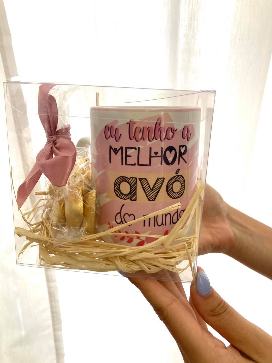 Caneca tenho a melhor avó do mundo Caneca com texto, saco dourado e laço rosa dentro de embalagem transparente, segurada por mãos com unhas azuis.