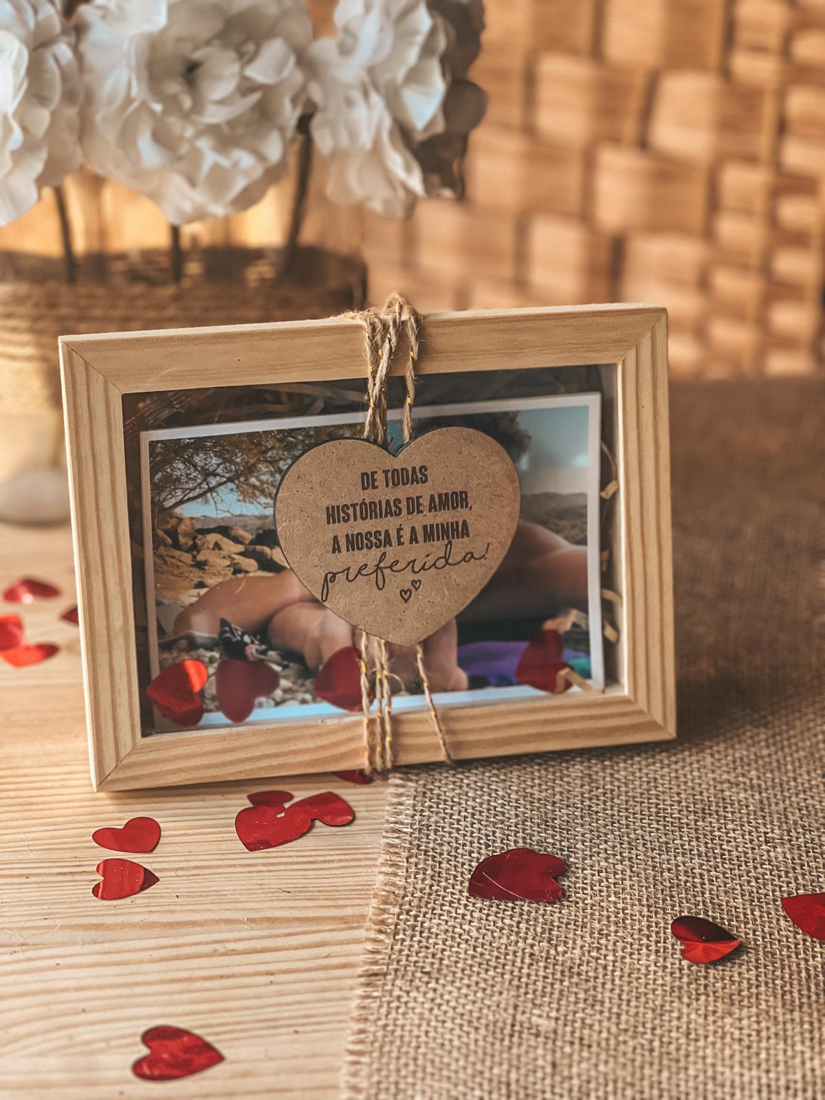 Caixinha com fotos e iman. Porta-retratos de madeira com pendente de coração e texto, confetes vermelhos, toalha de juta e flores ao fundo