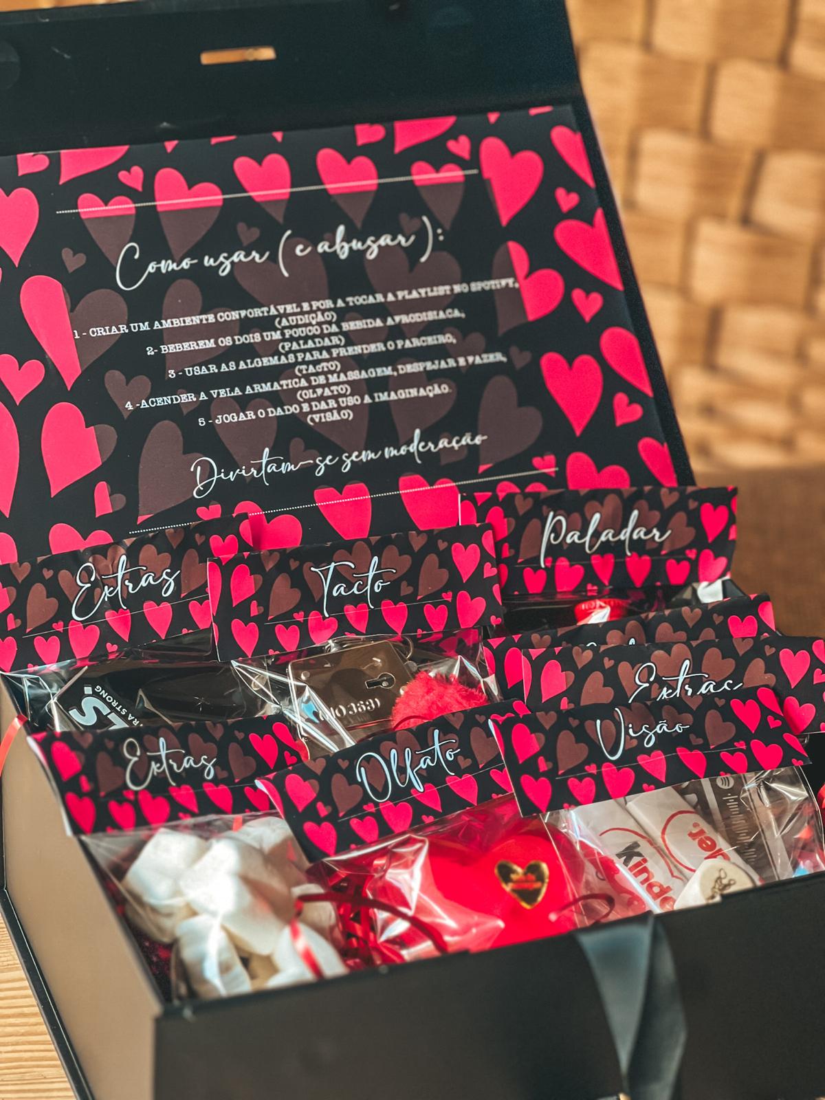 Caixa 5 sentidos Caixa preta aberta com pacotes decorados com corações e etiquetas com nomes dos sentidos.