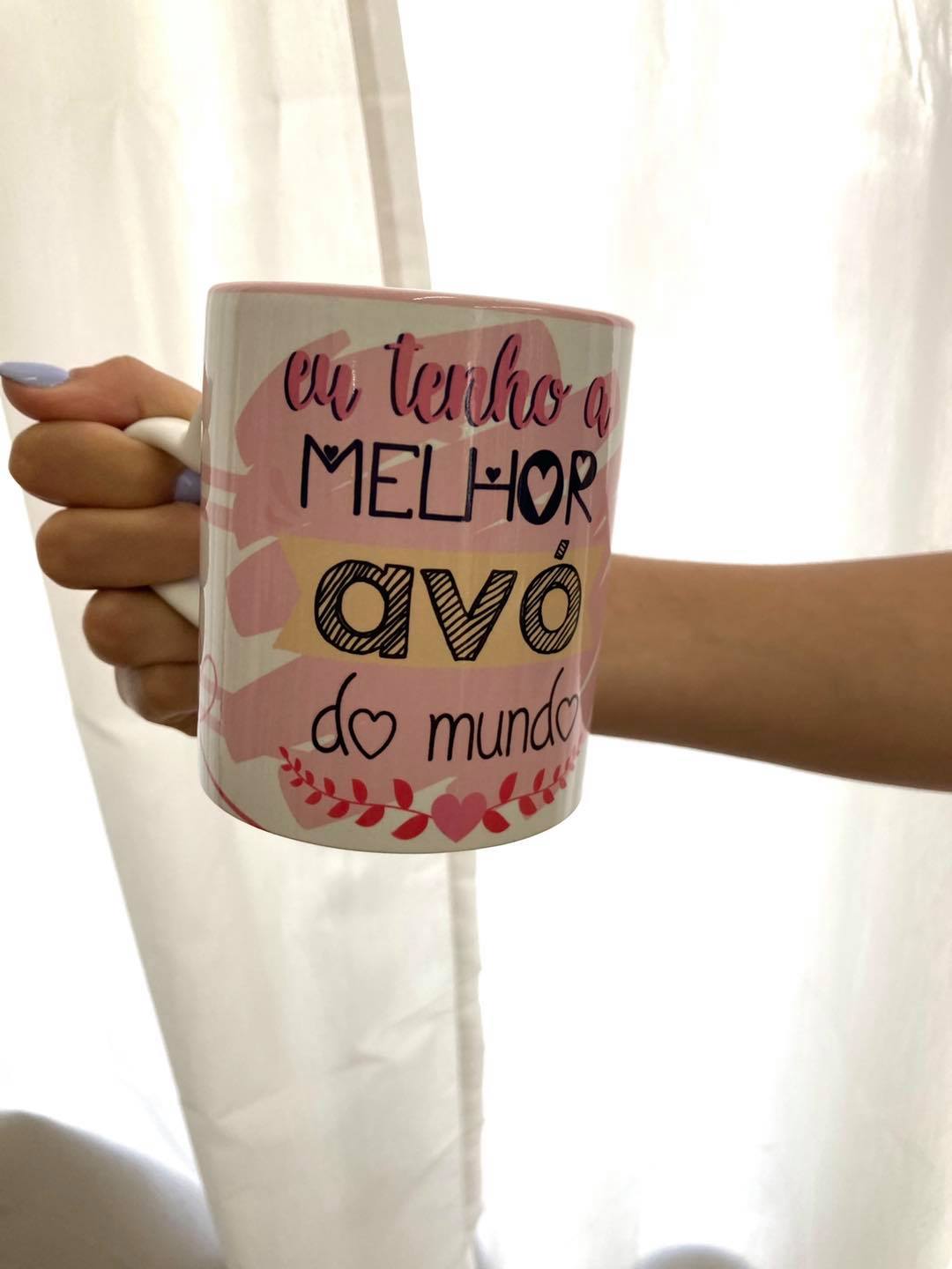 Caneca tenho a melhor avó do mundo Caneca branca com mensagem para avó, padrão floral rosa, segurada por uma mão