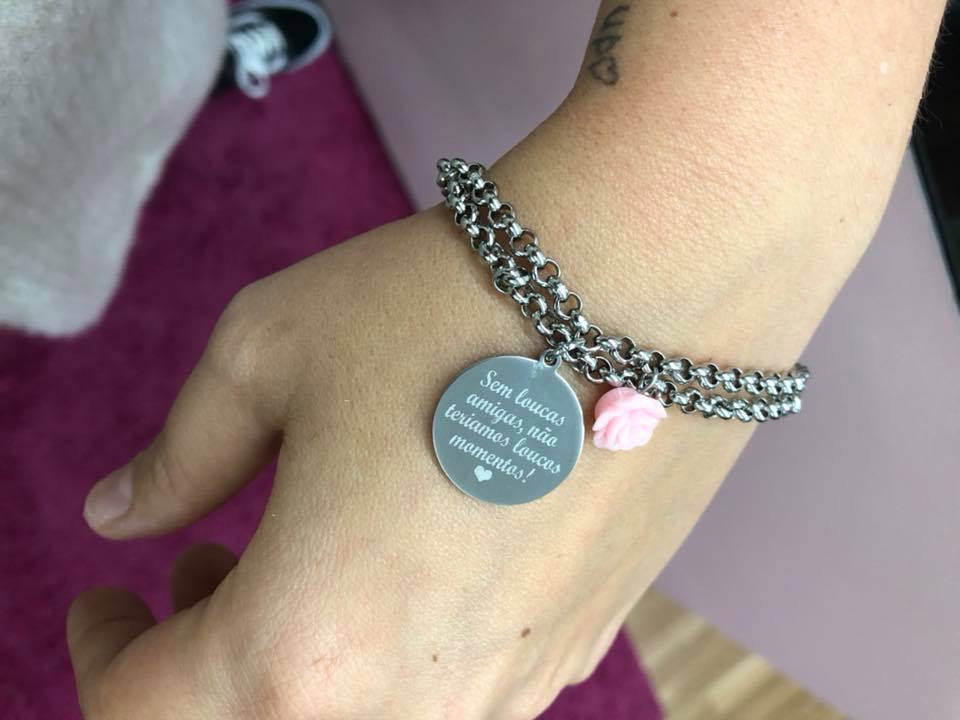 Pulseira sem loucas amigas Pulseira metálica prateada com pendente redondo cinza com texto em português e pequena flor rosa num pulso