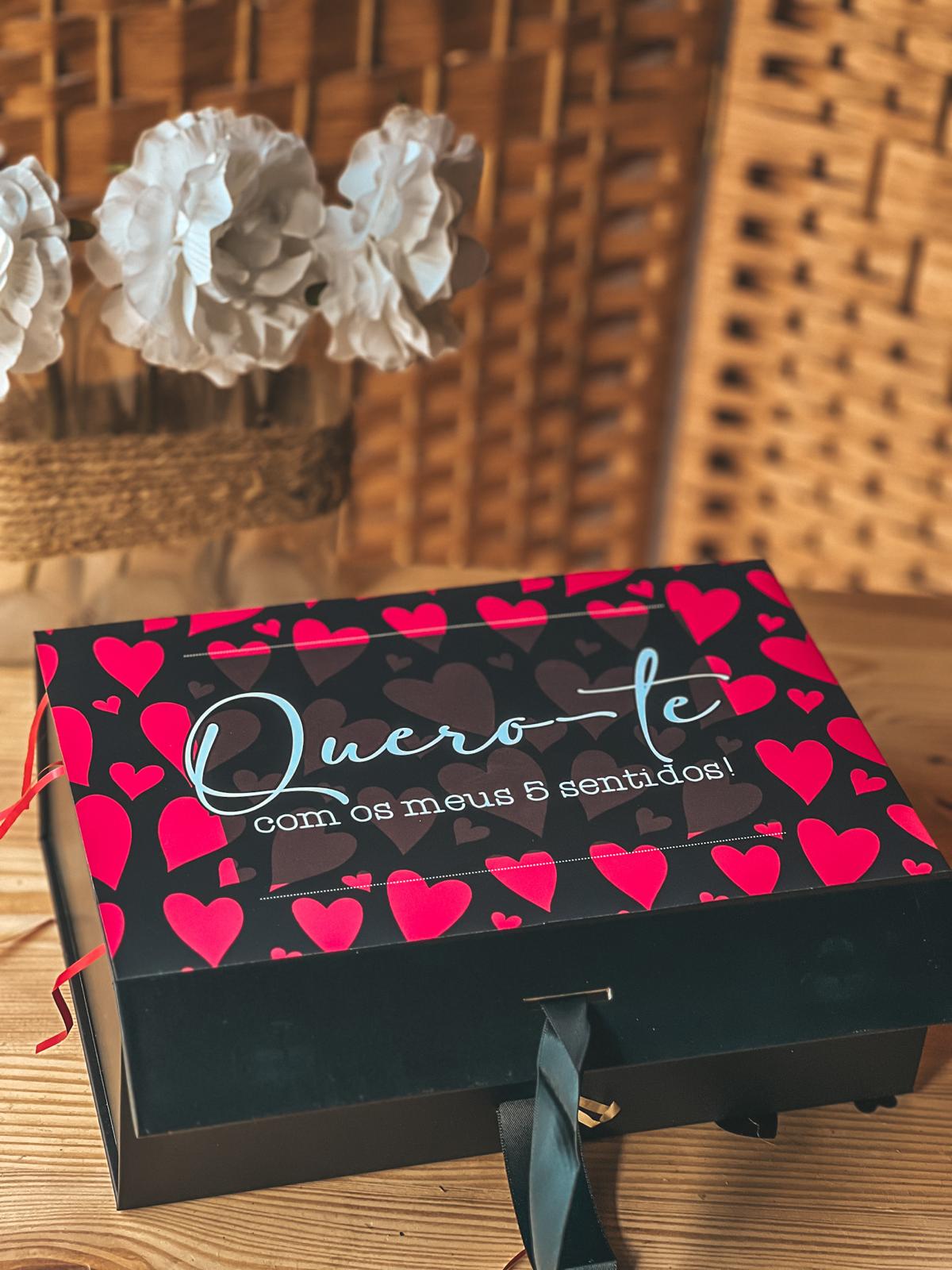 Caixa 5 sentidos Caixa de presente preta com corações vermelhos e frase romântica colocada sobre mesa de madeira com fundo em vime.