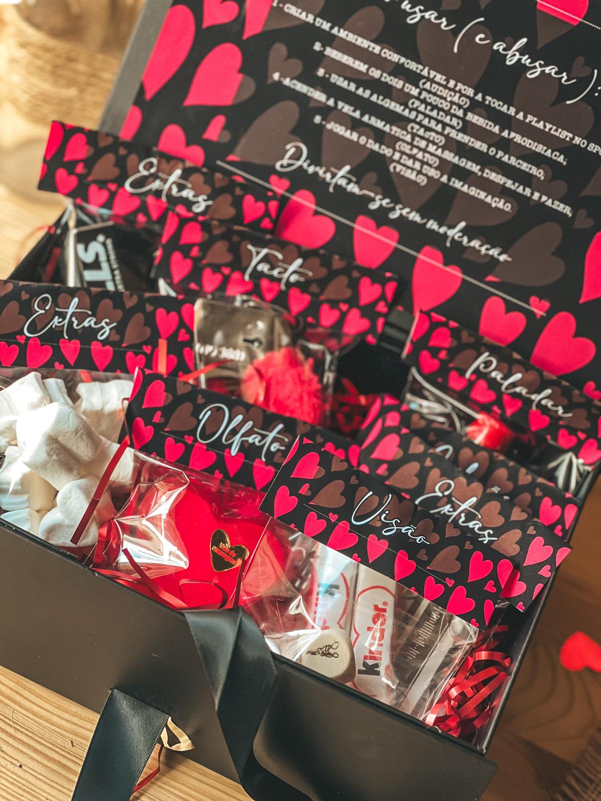 Caixa 5 sentidos Caixa presente preta com sacos transparentes com etiquetas de corações vermelhos e castanhos e doces variados no interior.