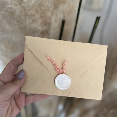 Envelope bege com selo de cera branco e cordão rosa sendo segurado por mão
