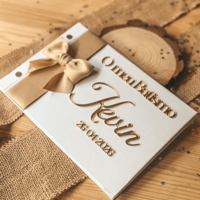 Livro de batismo personalizado branco com laço bege e letras douradas em relevo sobre mesa de madeira.
