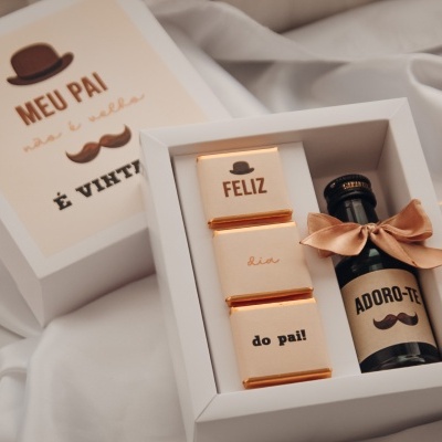 Caixa branca com chocolates dourados e garrafa preta com etiqueta 'ADORO-TE' e laço castanho
